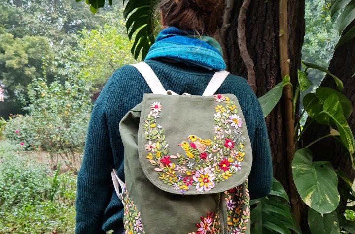 Seidenbandstickerei grüner Rucksack Vogel Person mit Rucksack voller Blumenstickerei, steht vor grüner Landschaft.